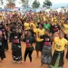 More than 100 Black women wearing different-colored "V20 1 Billion Rising" T-Shirts dance with their arms in the air on a reddish-dirt ground.
