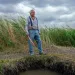 Jim Enote wears jeans and suspenders and stands atop a spring surrounded by tall grass. He's holding a long wooden instrument.