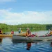 Four adults and two children travel in two canoes across a calm lake with trees in the background