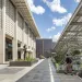 Students walk on a pathway next to a 1950/60s concrete building. To the right is a newly built outdoor area protected by panels that may be solar.