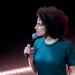 Ilana Glazer is on stage, holding a mic and looking to her right. She's wearing cut-off jean shorts and a teal T-shirt.