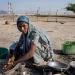 A woman wearing a scarf and Indian clothing washes dishes in a bowl outside. The Tata Mundra power pl​ant is in the background.