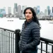 Varshini Prakash, wearing a dark-blue puffy jacket, looks into the camera while standing against a metal fence. Behind her is water and a city skyline.