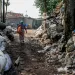 The back of midwife Mak Muji walking down a path toward a building in between two large piles of garbage and trees. In a bag, she's carrying the analog scale she uses to weigh babies.