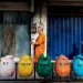 In Indonesia, three barefoot monks wearing saffron robes walk in a line, carrying silver pots for collecting alms. They're passing five bins shaped like tulips, which say (left to right) B3, Plastik, Organik, [illegible], and Kertas.