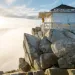 A hiker watches the sunrise from the porch of the Mt. Pilchuck fire lookout in Washington. The building is a white weathered cabin atop boulders.