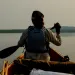 The back of a woman and a paddle at the front of a canoe on a calm body of water. The canoe has various bags and paddles stored in the middle.