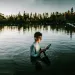 A young man stands in Lake Temescal, Oakland, at dusk holding an iPad. There's a soft glow on his face.