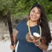 Moñeka De Oro smiles and stands on a beach holding a cut coconut and a machete.
