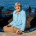 Ellen Shively sits on a rock by the ocean. Behind her are several seals.