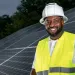 Jason Carney wears a hard hat and stands in front of a large solar array.