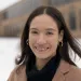 Alexandra Collins is smiling at the camera, wearing a brown coat and black turtleneck and standing in front of her high school with snow on the ground.