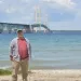 David Holtz stands on a beach with the Mackinac Bridge in the background. He's wearing a baseball cap, an unbuttoned shirt, and khakis.