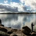 Two people are fishing from the shore of Millerton Lake. The water is glassy, and blue sky pokes through clouds above.