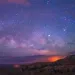Image of an indigo pinkish sky with a setting or rising sun in the background, and visible stars and galaxies. 