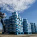 A woman in ripped jeans and a windbreaker hands a package of water to a person not in the photo. A pallet of bottled water is behind her.