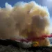 Cloud of fire retardent bursting over a green-brown hilly landscape that is on fire, against a blue sky.
