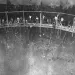 Black and white photo of tiny people in workwear on a narrow walkway inside a massive metal cavern of some kind.