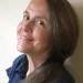 Naomi Shihab Nye looks to the left over her shoulder and smiles at the camera. Nye has long, dark hair and is wearing a dark-blue button-up shirt.