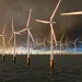 Row of wind turbnes standing in a darik ocean, against a moody sky against a setting sun.