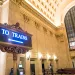 Massive cream-colored stone room in Chicago's Union Station, with an illuminated sign of a blue arrow on a black background reading "to trains."