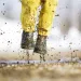 Child photographed from the waist down jumping into a puddle with yellow pants and black rain boots