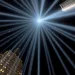 Skyscrapers against a night sky as seen from below, with spotlights on all sides meeting in the center