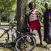 Two women wearing summer clothes pose in front of a tree in a park, smiling. A bike is leaned up against the tree.