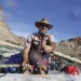 A man steers a boat on a river with two oars. Behind him is blue sky and desert/rock walls.