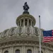 Top of the US Capitol building, with an American flag flying out front and a cloudy sky overhead.