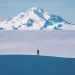 A hiker walks across a snowy ridge with a layer of fog and Glacier Peak in the distance.