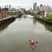 A red kayak floats down a brown, murky canal with industrial buildings along the sides and tall city buildings in the distance.