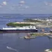 Aerial photo shows a large blue and white ship leaving a port. Onshore are five short white cylinders and other infrastructure.
