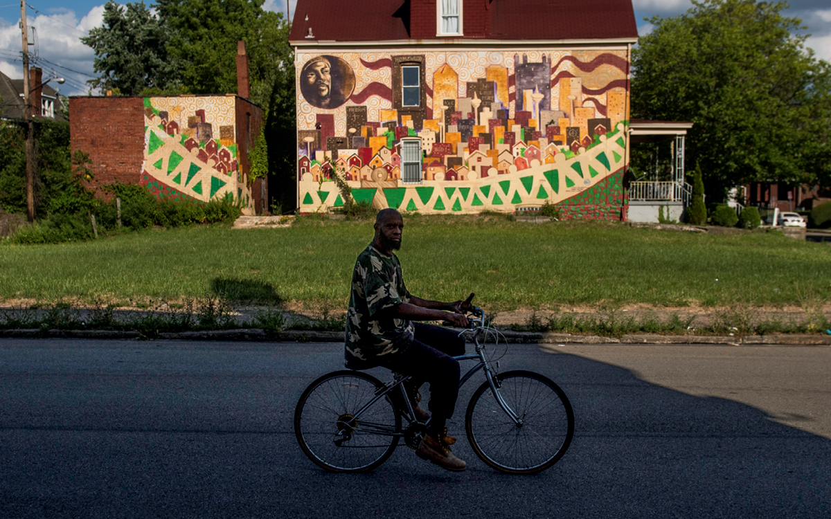 A man bicycles down Homewood Avenue. Behind him, a colorful mural is painted on the side of a house.