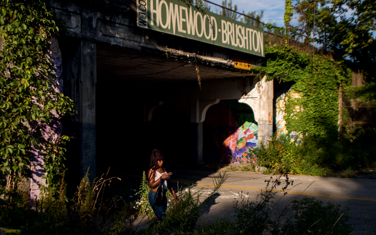 A woman walks under an overpass. Above her, a large sign reads "Welcome Homewood-Brushton."