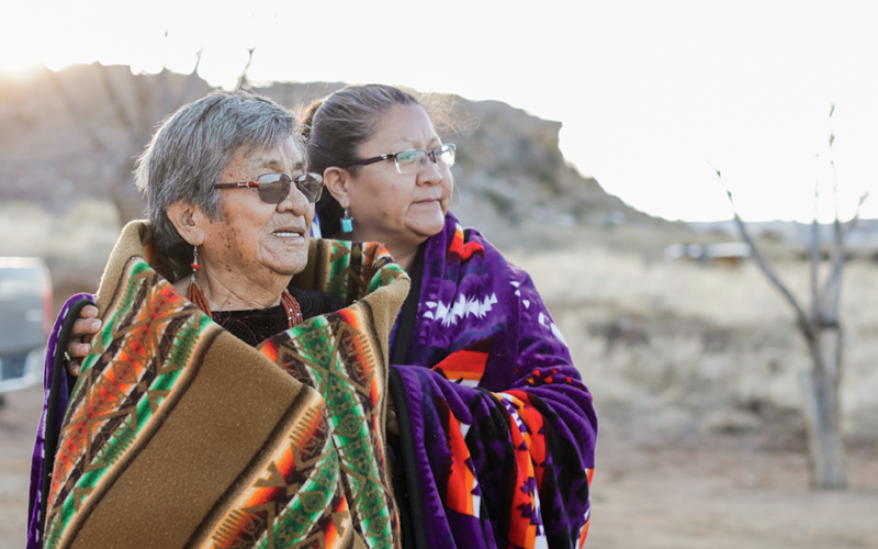 Jessie and Verma Harrison stand together, both wearing blankets around their shoulders and looking off in the distance.
