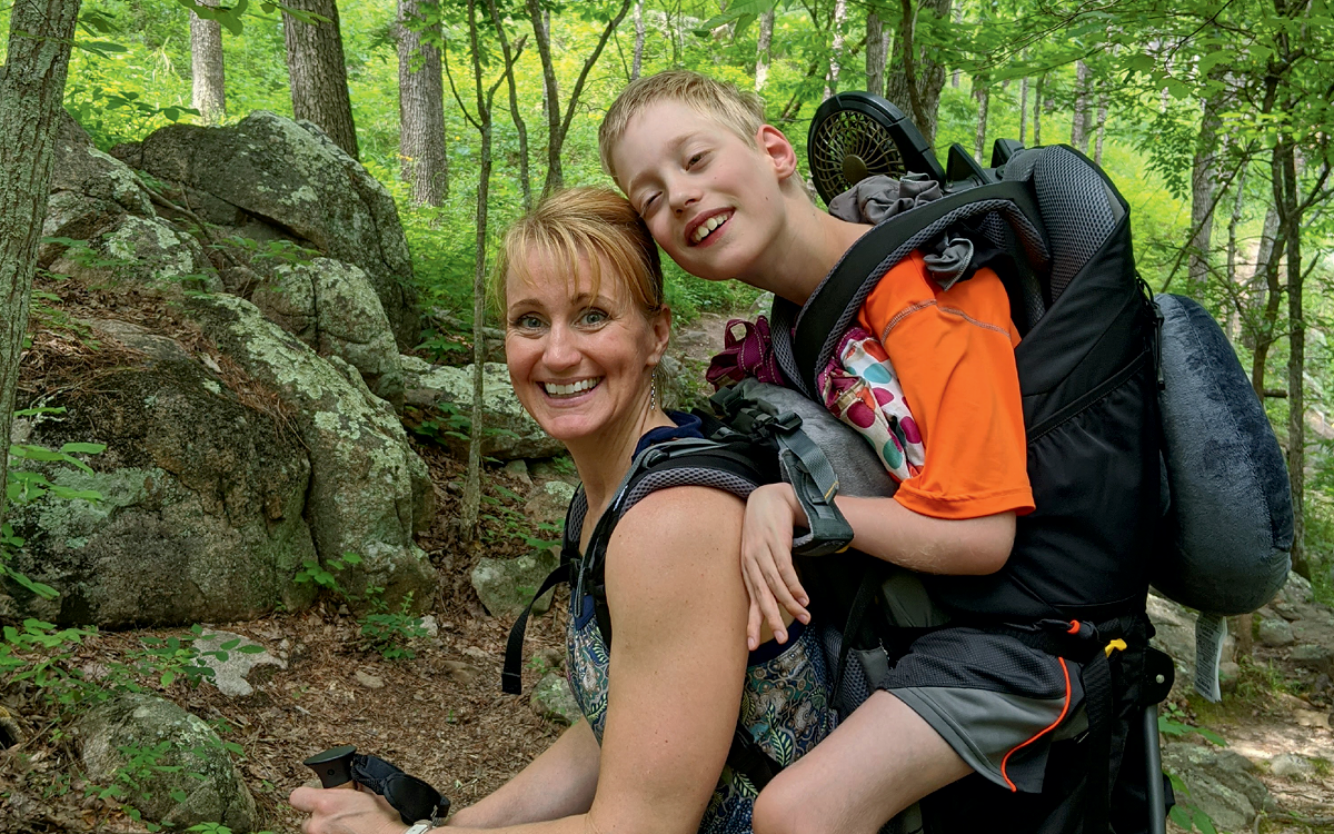 A woman hikes with a boy on her back in a WCK Pack. They're both smiling at the camera.
