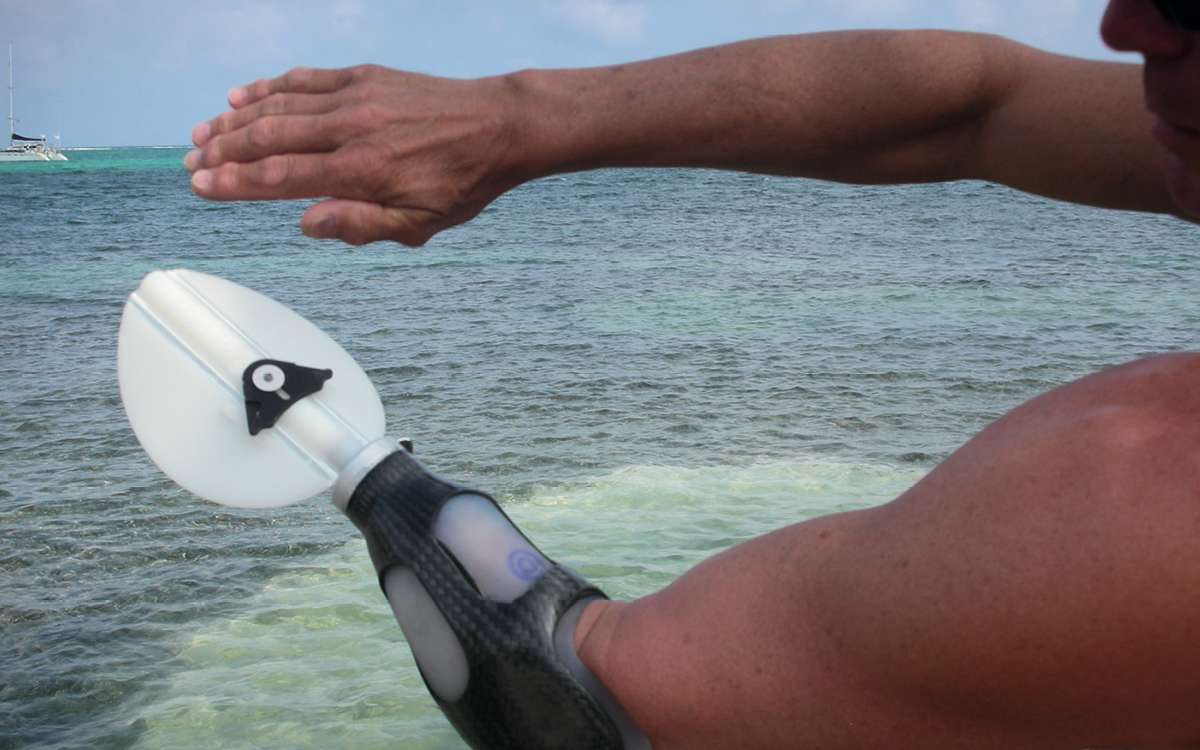 Someone holds out their arms in front of an ocean. One arm has a TRS Prosthetics Freestyle Swimming Device.