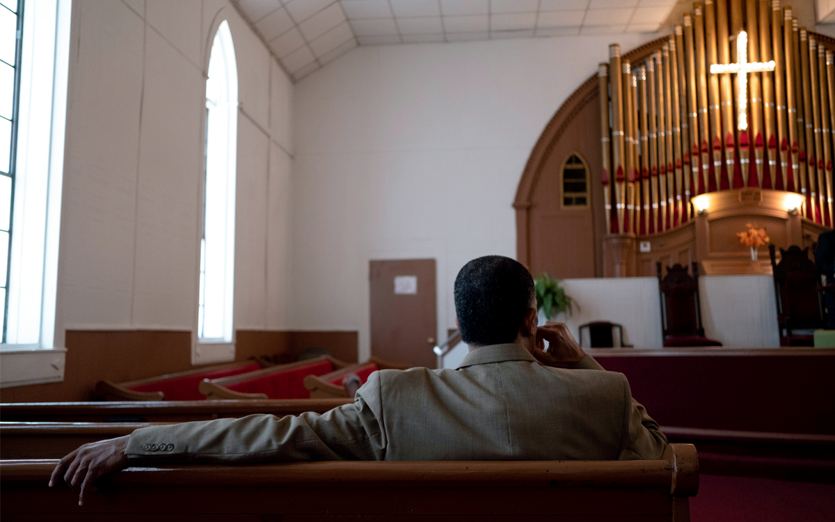 A man sits in a church pew with his arm draped over the back. Image shows him from behind as he looks up toward the stage.