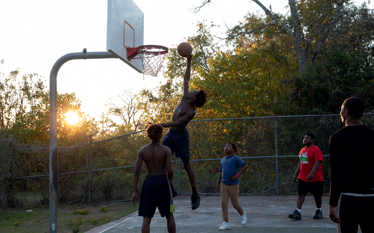 Five young men play basketball at sunset. One of them is jumping up for a dunk.