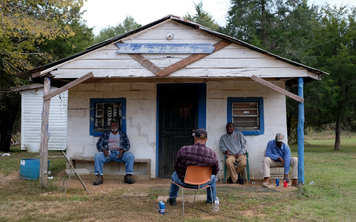 Four men sit and drink cans of beer outside a dilapidated building with "Bright Eyes Places" handpainted in cursive on a sign overhead.