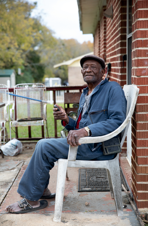 Deacon Samuel Woods sits in a white plastic chair outside a brick building and looks at the camera.