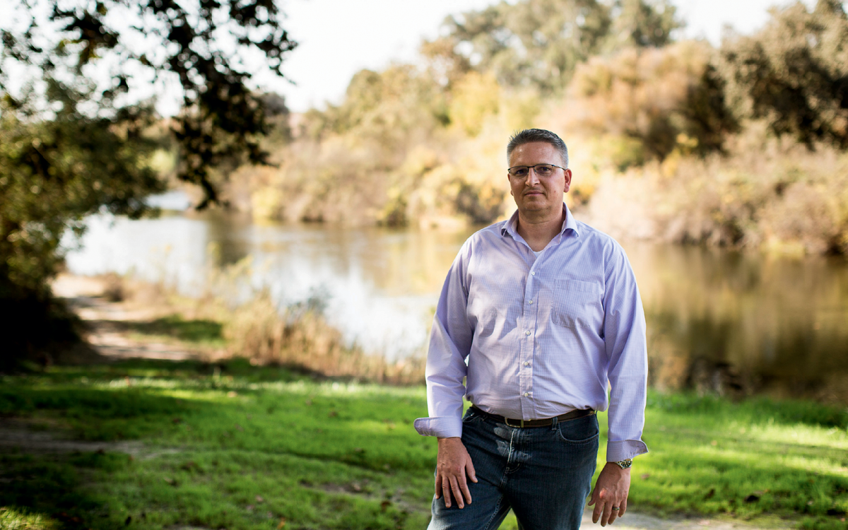 Don Portz, wearing glasses and a buttoned-up shirt, stands in front of a river and looks at the camera.
