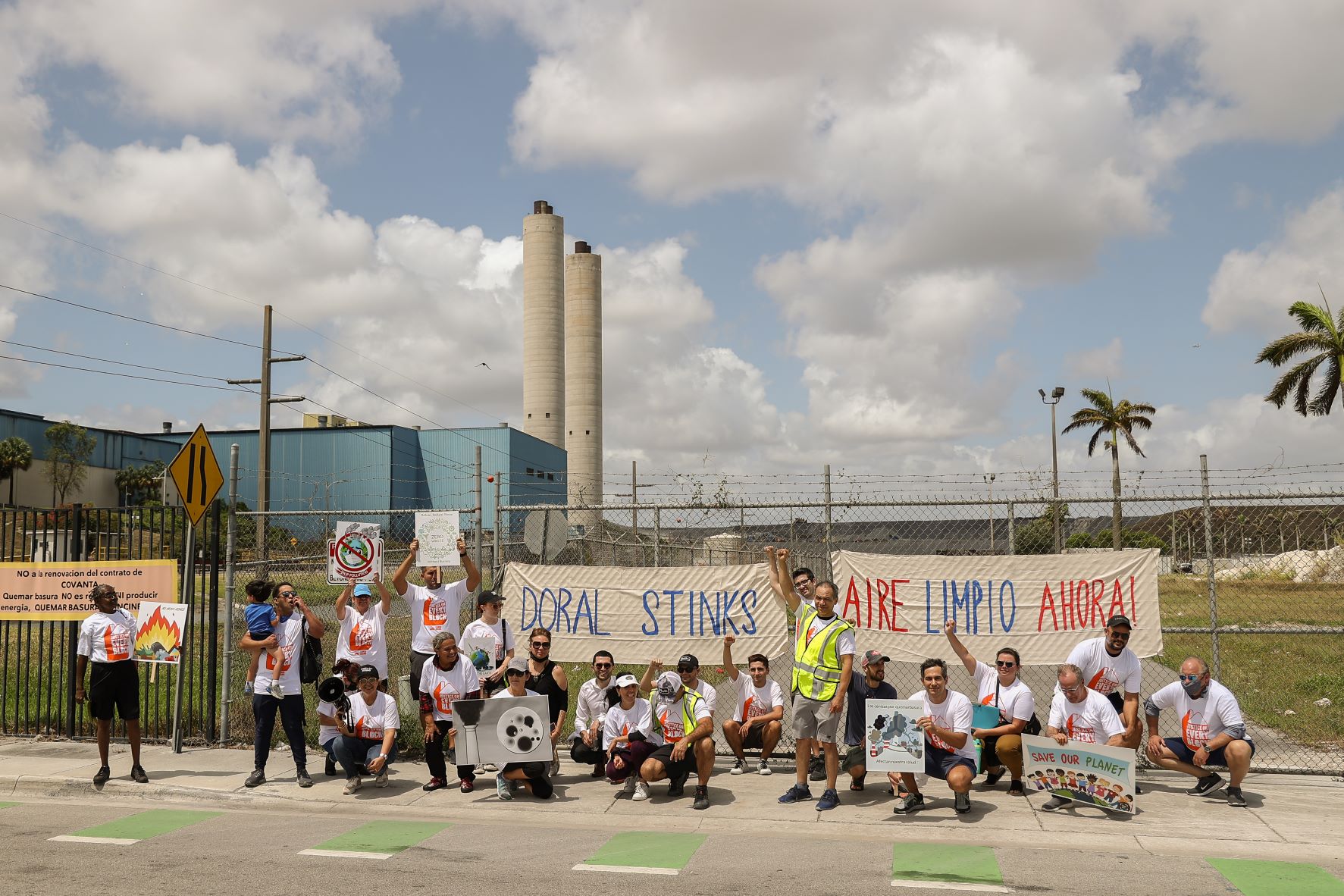 In front of smokestacks, banners reading "Aire Limpio Ahora!" and "Doral Stinks!", along with a crowd of activists