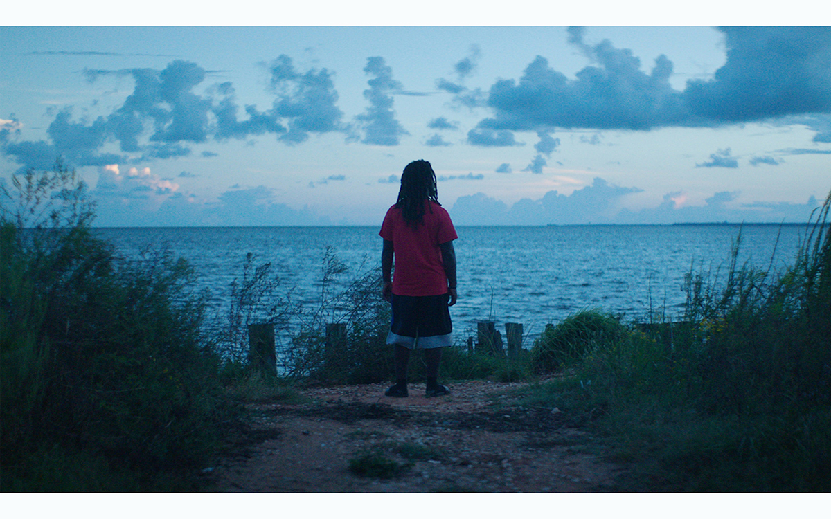 A person with long dreadlocks and long atheltic shorts stands at the edge of an ocean at dusk, looking into the blue distance.