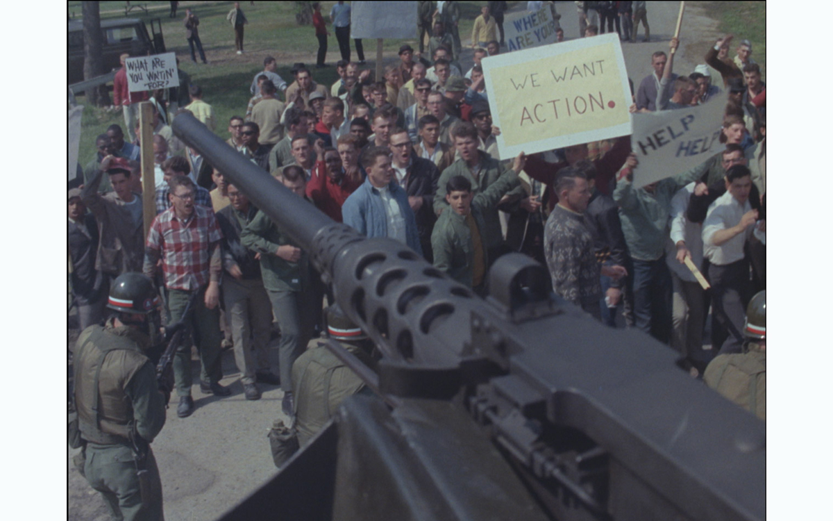 A 1960s era tank gun turret points diagonally over a crowd of army recruits dressed as 1960s era protestors, complete with signs
