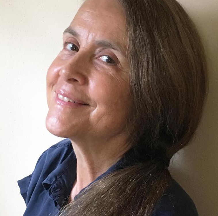 Naomi Shihab Nye looks to the left over her shoulder and smiles at the camera. Nye has long, dark hair and is wearing a dark-blue button-up shirt.