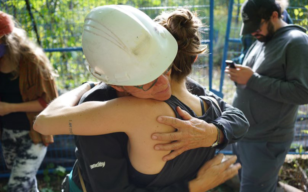 Two figures embracing in the forest. One is wearing a hard hat and blue jump suit, and the other is in a tank top. 