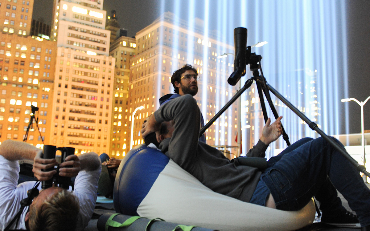 People lying on the roof of a parking garage next to beams of light, looking for birds in the sky