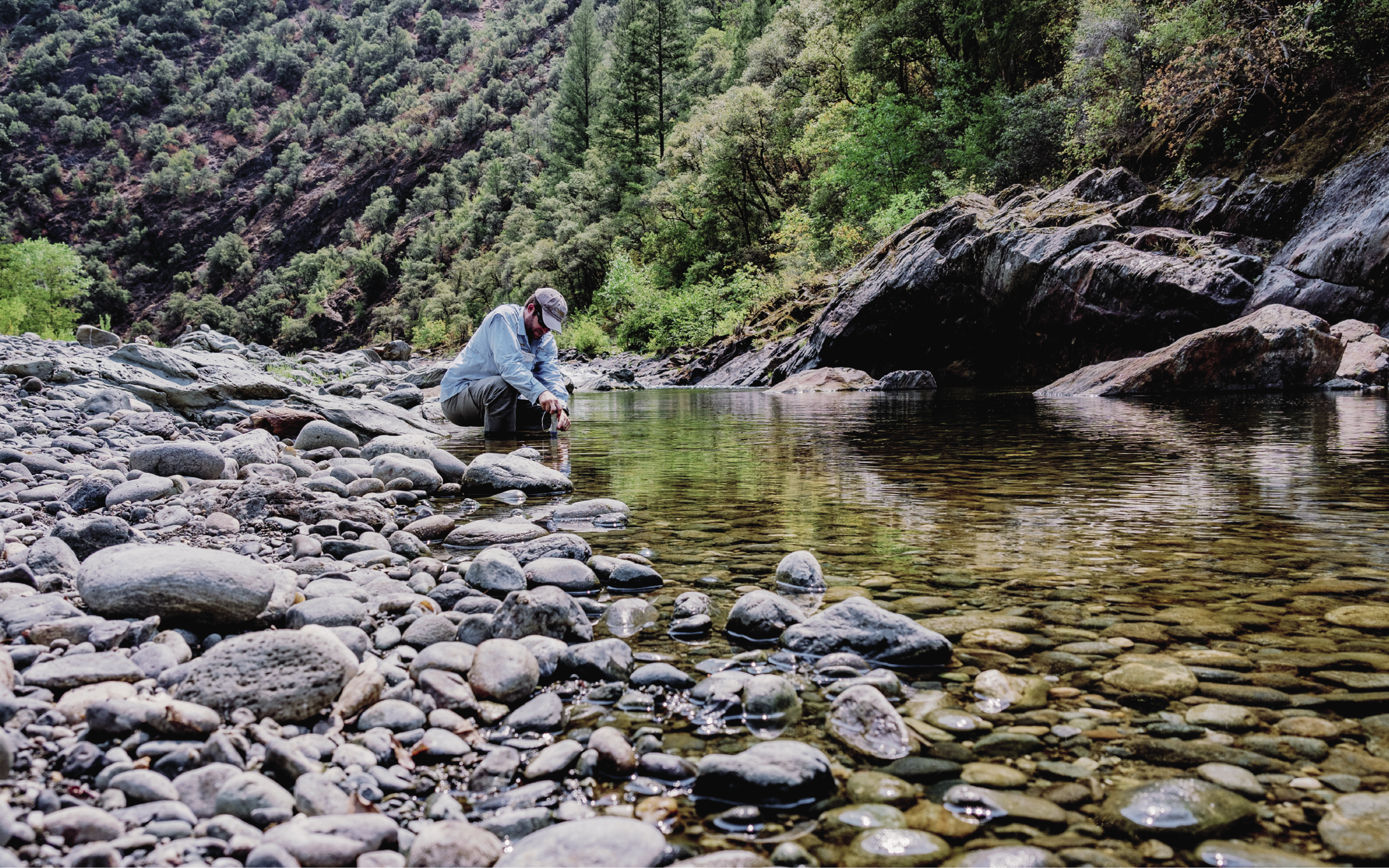 Ryan Peek is wearing a blue shirt and kneeling down next to a rocky river.